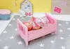 A baby doll is lying in a pink baby bed on a star-patterned rug. Produktbild-Vorschau