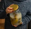 A person holding a large glass jar filled with a cloudy liquid and green objects, with the gold lid partially removed. Podgląd zdjęcia produktu