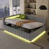 A modern bedroom with a grey upholstered bed featuring integrated LED lighting and storage. The bed is made up with a white mattress, a brown throw blanket, and a green decorative pillow. The room has large windows with a city view and shelves with decorat Produktbild-Vorschau