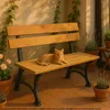 An orange cat is resting on a wooden park bench on a tiled patio, with potted plants and ivy in the background. Produktbild-Vorschau