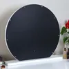 The back of a round, white-framed mirror with a black backing, resting on a white stand on a white surface. A red flower in a vase and some cosmetic items are visible on the right. Náhled obrázku produktu