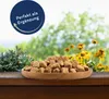 A wooden bowl filled with brown cubes of food, placed on a wooden surface with a blurred background of green foliage and yellow flowers. Produktbild-Vorschau