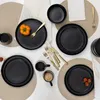 A top-down view of a black ceramic dinnerware set arranged on neutral placemats, with decorative elements like a small orange pepper and cherry tomatoes. Voir les images du produit