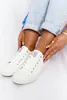 A pair of white low-top sneakers with white laces and a white sole, worn by a person sitting down. The person is wearing light blue jeans and has several rings on their fingers and an anklet. Podgląd zdjęcia produktu