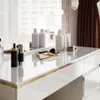A vanity table with various cosmetic products arranged on its surface, with a blurred background of a window and curtains. Produktbild-Vorschau