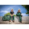 Two children are sitting on a stone wall outdoors, wearing green overalls and caps. Produktbild-Vorschau