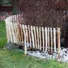A rustic wooden fence made of natural hazelnut branches is installed in a garden, bordering a bush and grass. Produktbild-Vorschau