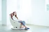 A woman is relaxing in a massage chair in a bright, minimalist room. Produktbild-Vorschau
