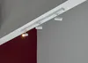 A white track lighting system with three adjustable spotlights mounted on a white track attached to a ceiling. The background consists of a gray wall and a dark red wall. Náhľad obrázku produktu