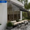 An outdoor seating area with a black awning extended over a window, featuring a bench with cushions and tables with chairs. Náhľad obrázku produktu