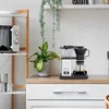 A coffee maker on a kitchen counter next to a plant and a microwave. Produktbild-Vorschau