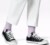 A close-up shot of a person's feet wearing black Converse sneakers and light purple socks, with white pants. Voir les images du produit