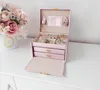 A pink jewelry box with gold accents is open on a white dresser, next to a vase of pink flowers. Náhled obrázku produktu