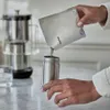 A person is pouring coffee grounds from a pouch into a stainless steel coffee maker. Anteprima dell’immagine prodotto