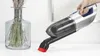 A hand is holding a cordless vacuum cleaner, using it to clean up small debris on a white surface next to a glass vase with dried flowers. Voir les images du produit