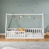 A white wooden house bed for children, set up in a room with a light blue wall decorated with star decals. A string of colorful pom-pom lights hangs above the bed. Produktbild-Vorschau