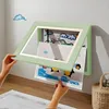 A person is holding a child's artwork in a light green frame that is open and attached to a wall. There is a cloud sticker on the wall. Produktbild-Vorschau