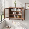A wooden wall-mounted cabinet with glass doors, filled with various bathroom products, is shown in a well-lit room with a window. Produktbild-Vorschau
