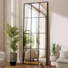 A tall, rectangular mirror with a black metal frame, resembling a window pane, is placed in a well-lit room. It reflects a living space with curtains and furniture. Two potted plants, one large palm and one smaller snake plant, flank the mirror. A portion  Produktbild-Vorschau