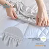 Hands are shown stretching a light gray fitted sheet over a white quilted mattress pad. Produktbild-Vorschau