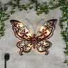 A decorative butterfly wall light with colorful jewel accents, mounted on a textured gray wall, with ivy vines framing the top. Produktbild-Vorschau
