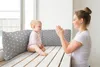 A mother and baby are sitting on a wooden bench by a window, with pillows behind them. The mother is clapping her hands, and the baby is looking at her. Produktbild-Vorschau