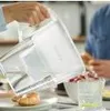A person is pouring water from a pitcher into a bowl of red berries. There is a croissant on a plate in the foreground. Produktbild-Vorschau