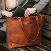 A person is putting a laptop into a brown leather tote bag while sitting on a wooden bench. Voir les images du produit