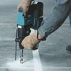 A person wearing gloves is using a Makita rotary hammer drill to drill into a concrete floor. Produktbild-Vorschau