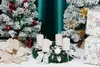 A Christmas-themed arrangement with two small, snow-dusted artificial trees, an advent wreath with four white candles, a large white poinsettia decoration, and wrapped gifts. Anteprima dell’immagine prodotto