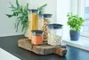 A collection of clear storage containers filled with dry goods, arranged on a wooden cutting board, with plants in the background. Produktbild-Vorschau