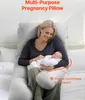 A woman is holding a baby while sitting on a bed with a large U-shaped pregnancy pillow. Produktbild-Vorschau