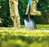 A person's legs and feet wearing work boots and tan pants are shown, with one foot stepping on a shovel blade. The shovel is partially inserted into a grassy lawn. Lush green bushes are visible in the background. Náhľad obrázku produktu