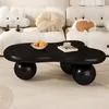 A black coffee table with a unique, organic shape sits on a cream-colored rug in front of a white sofa. The table has a bowl of fruit and a book with a cup on it. Decorative pillows, including a cloud-shaped one and a round one, are on the sofa. Voir les images du produit
