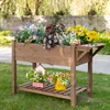 A wooden raised garden bed filled with artificial flowers, placed outdoors on grass with a stone wall and trees in the background. Produktbild-Vorschau