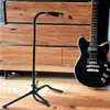 A black guitar stand and a black electric guitar are placed on a wooden floor in front of a wooden dresser. Náhľad obrázku produktu