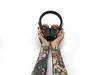 A person's tattooed arms are holding a pair of black headphones above their head against a white background. Voir les images du produit
