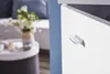 A close-up of a white cabinet with a silver handle, with a blurred background of a living room setting. Náhled obrázku produktu