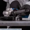 A person wearing black gloves is holding a cordless angle grinder over a metal surface. Produktbild-Vorschau