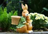 A decorative statue of two rabbits in a garden setting with flowers and greenery. Produktbild-Vorschau