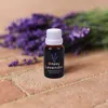 A bottle of lavender fragrance oil is placed in front of a bunch of lavender flowers, on a brick-like surface. Produktbild-Vorschau