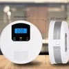 Two white carbon monoxide detectors are shown on a wooden surface. One is shown from the front, displaying a digital readout and indicator lights. The other is shown from the side, highlighting its ventilation grilles. Dimensions are indicated with lines a Produktbild-Vorschau