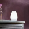 A white, egg-shaped LED light sits on a gray surface in front of a blurred purple background. A glass vase with pink flowers is visible to the left. Produktbild-Vorschau