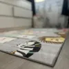 A close-up, low-angle shot of a children's rug with animal designs on a wooden floor. A blurred dresser is visible in the background. Produktbild-Vorschau