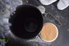 A black ceramic storage jar with a wooden lid and a tea infuser on a dark, sprinkled surface. Produktbild-Vorschau