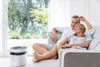 A couple relaxing on a couch in a bright living room with an air purifier in the foreground. Produktbild-Vorschau