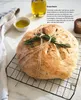 A loaf of bread with rosemary on a cooling rack, next to a small bowl of olive oil and a glass of olive oil. Produktbild-Vorschau