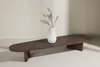 A dark brown, oval-shaped coffee table with a white vase containing dried plants on top, set against a light-colored wall. Produktbild-Vorschau
