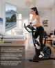 A woman is using a stationary exercise bike in a living room setting, with a large screen displaying a scenic road in the background. Produktbild-Vorschau