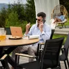 A man is working on his laptop and talking on the phone at a table outdoors, with a woman relaxing in a hanging chair in the background. Produktbild-Vorschau